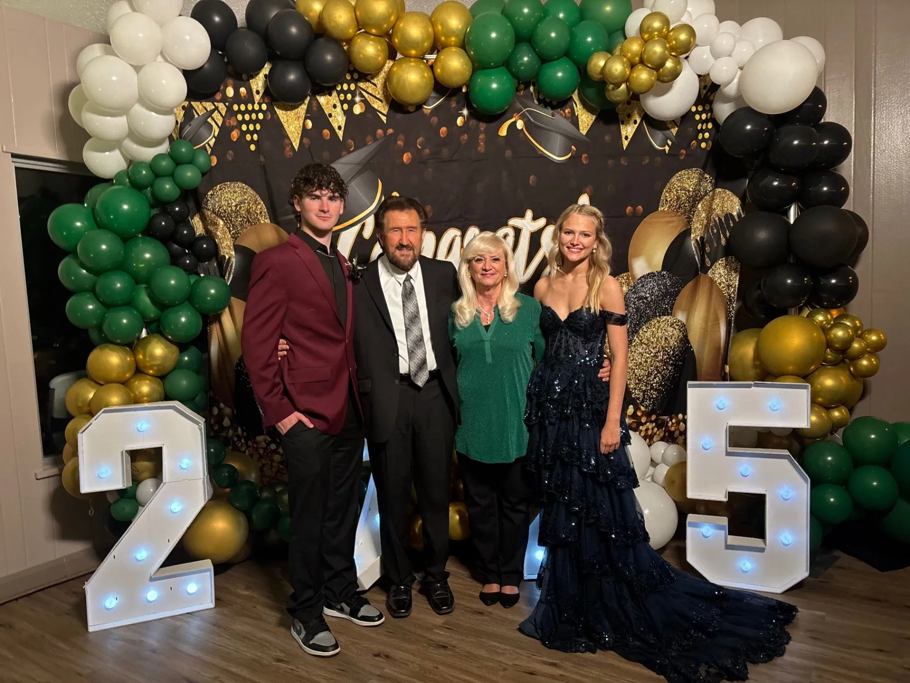 Graduation celebration with a green, gold, black, and white balloon garland, themed backdrop, and illuminated 25 marquee numbers