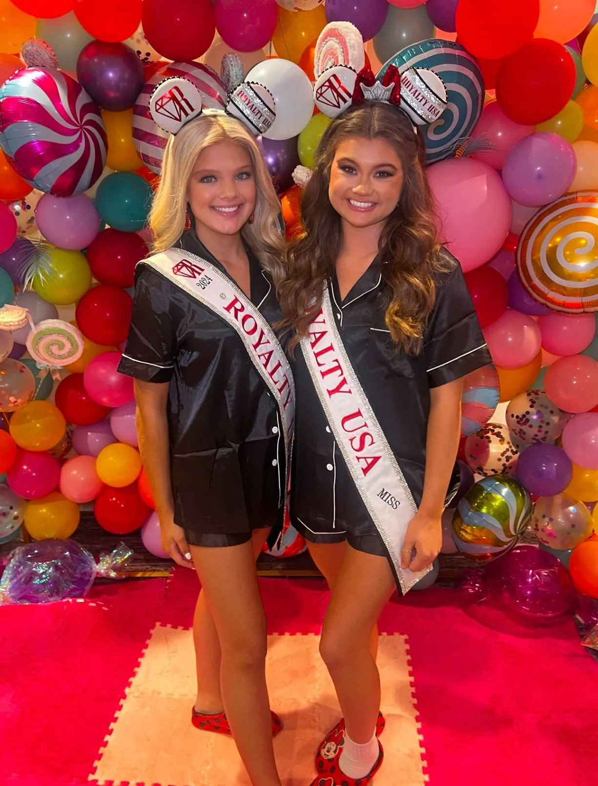 Two titleholders in silk pajamas in front of a bright candy-colored balloon wall